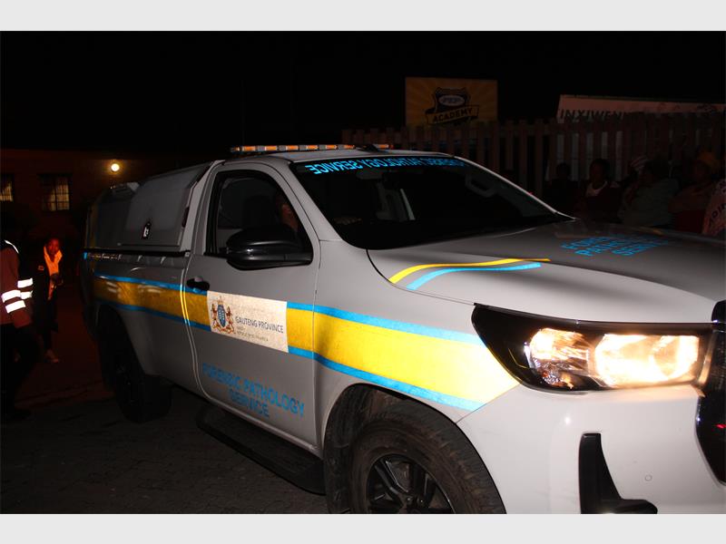 A white police vehicle with bright blue and yellow stripes is parked at night, its headlights are on. People and buildings are dimly visible in the background.