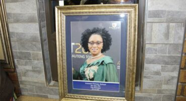 Framed portrait of a person in a green gown with a sash, set against a backdrop with text, placed on a stone mantel. Formal and celebratory tone.