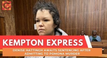 A person in a gray hoodie sits in a courtroom, looking serious. The wood-paneled walls and benches create a formal atmosphere. Two people sit in the background.