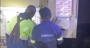 Two utility workers are examining an open electrical panel at night. They are wearing high-visibility jackets. A safety diagram is visible inside the panel.