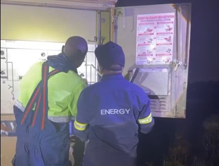 Two utility workers are examining an open electrical panel at night. They are wearing high-visibility jackets. A safety diagram is visible inside the panel.