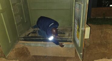 A worker in a blue jacket inspects cables inside an open utility box at night. The scene is dimly lit, highlighting the worker's task-focused posture.