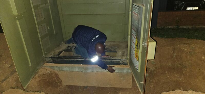 A worker in a blue jacket inspects cables inside an open utility box at night. The scene is dimly lit, highlighting the worker's task-focused posture.