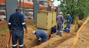 Construction workers in blue uniforms dig a trench beside a sidewalk, repairing underground utilities. A large electrical box is nearby, with trees in the background.
