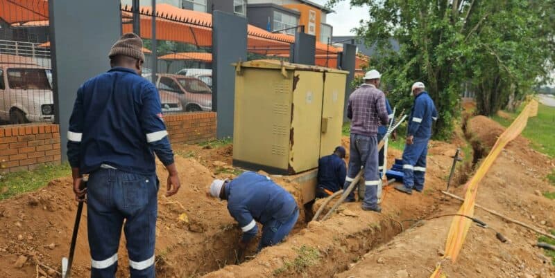 Construction workers in blue uniforms dig a trench beside a sidewalk, repairing underground utilities. A large electrical box is nearby, with trees in the background.