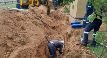 Workers in protective gear dig a trench near a roadside with a backhoe loader nearby. The scene is industrious and teamwork-focused.