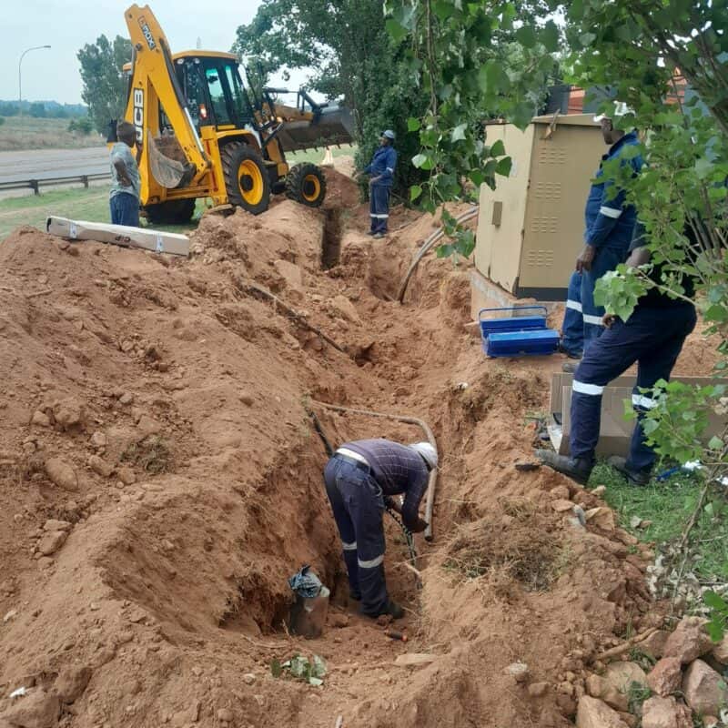 Workers in protective gear dig a trench near a roadside with a backhoe loader nearby. The scene is industrious and teamwork-focused.