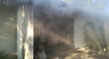 The image shows a house with a white exterior partially obscured by thick smoke emitting from a window. Sunlight filters through, creating an eerie atmosphere.