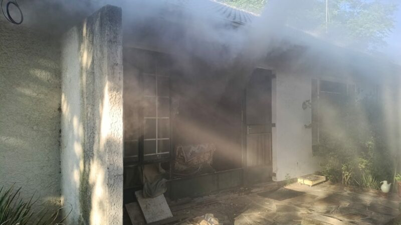 The image shows a house with a white exterior partially obscured by thick smoke emitting from a window. Sunlight filters through, creating an eerie atmosphere.
