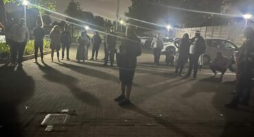 A group of people stand at night in a dimly lit parking lot, forming a loose circle. Streetlights cast long shadows, creating a calm, reflective atmosphere.