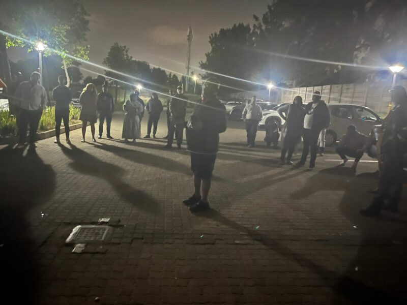 A group of people stand at night in a dimly lit parking lot, forming a loose circle. Streetlights cast long shadows, creating a calm, reflective atmosphere.