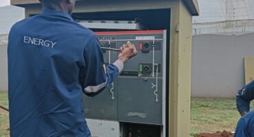 A worker in a blue "ENERGY" uniform and red cap operates a utility box outdoors under a clear sky, suggesting maintenance or electrical work.