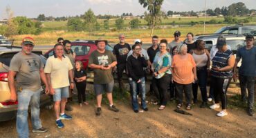 A diverse group of people stands outdoors on a dirt path next to parked cars. They appear relaxed and are gathered under a partly cloudy sky with a rural landscape in the background.