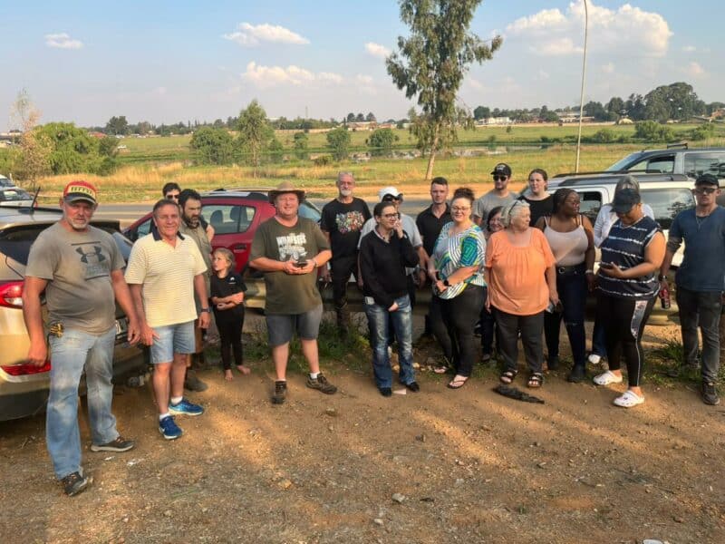 A diverse group of people stands outdoors on a dirt path next to parked cars. They appear relaxed and are gathered under a partly cloudy sky with a rural landscape in the background.