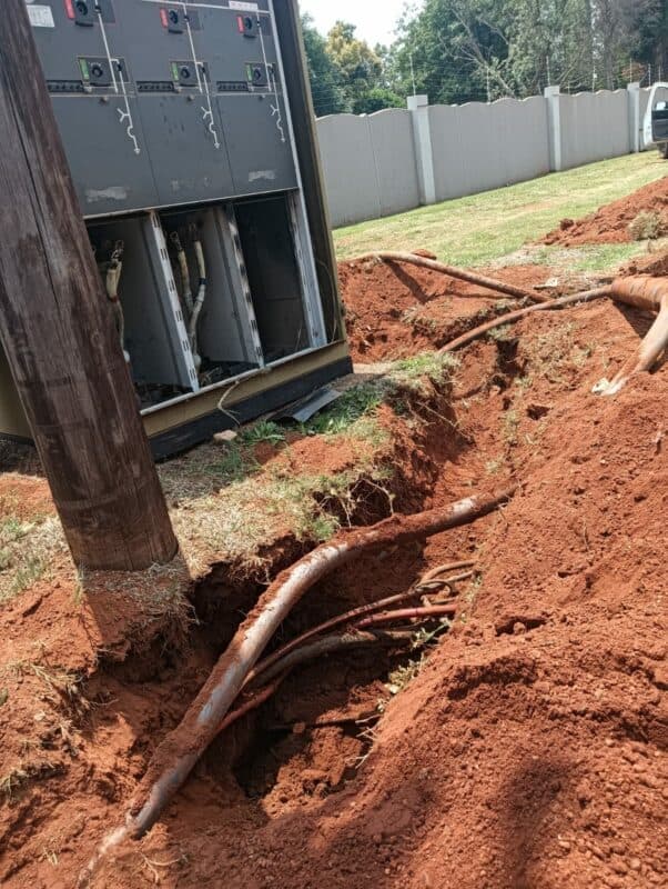 Underground cables exposed beside a utility box with open panels, surrounded by fresh red soil and dug trenches. The scene suggests ongoing repairs.