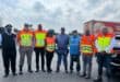 A group of eight people in safety uniforms and reflective vests stands smiling on a road under a partly cloudy sky, with a truck in the background.