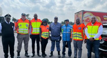 A group of eight people in safety uniforms and reflective vests stands smiling on a road under a partly cloudy sky, with a truck in the background.