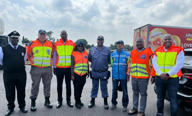 A group of eight people in safety uniforms and reflective vests stands smiling on a road under a partly cloudy sky, with a truck in the background.