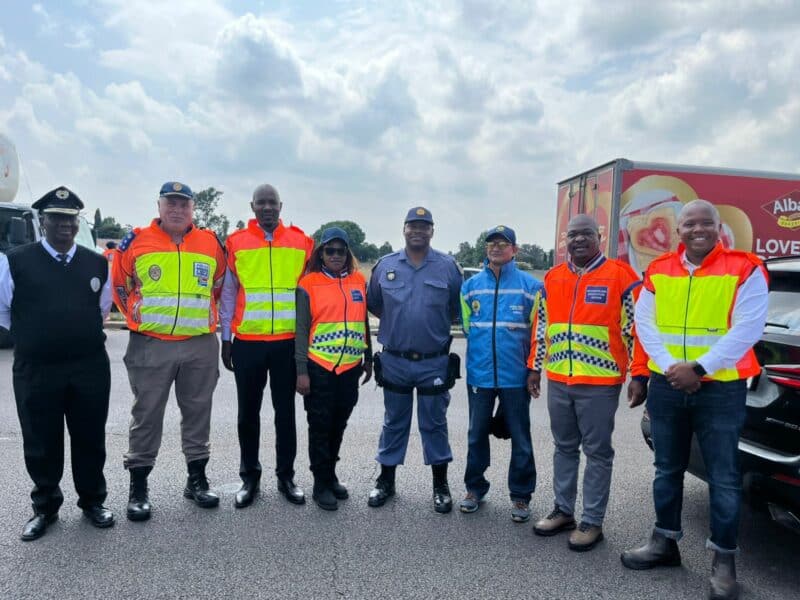 A group of eight people in safety uniforms and reflective vests stands smiling on a road under a partly cloudy sky, with a truck in the background.