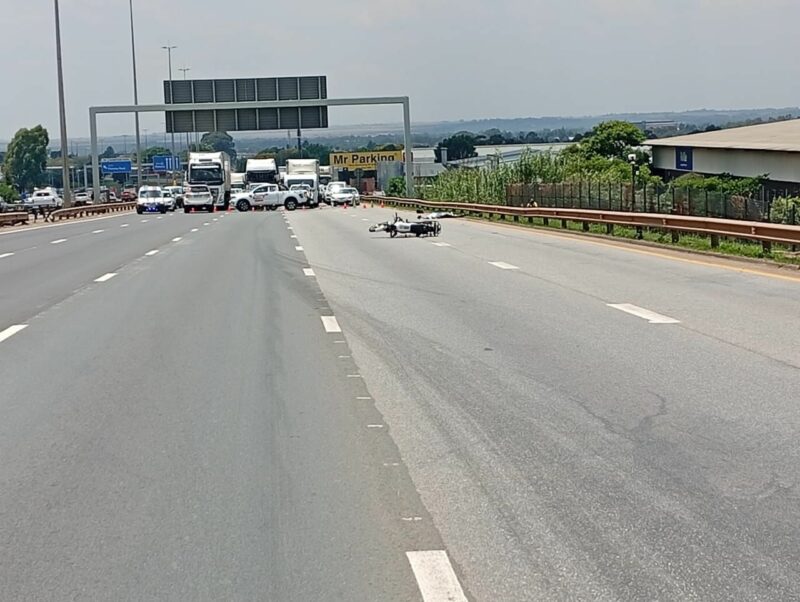 Wide highway with trucks and emergency vehicles blocking lanes ahead. A bike rests on the road surface. Overcast sky and a