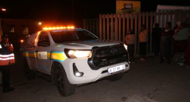 A police vehicle with flashing amber lights parked on a dimly lit street at night, surrounded by a group of people, conveying a tense atmosphere.
