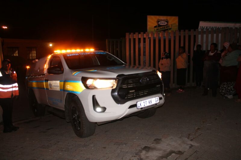 A police vehicle with flashing amber lights parked on a dimly lit street at night, surrounded by a group of people, conveying a tense atmosphere.