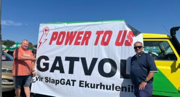 Two men stand smiling beside a large banner reading "Power to Us, Gatvol" on a sunny day. A yellow and green vehicle is partially visible in the background.
