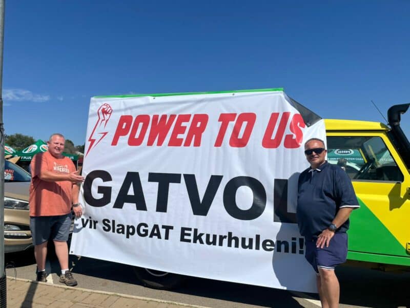 Two men stand smiling beside a large banner reading