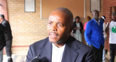 A man in a suit speaks into a microphone against a brick wall backdrop. People stand in the background, some wearing blue and white clothing.