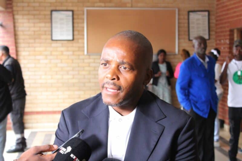 A man in a suit speaks into a microphone against a brick wall backdrop. People stand in the background, some wearing blue and white clothing.