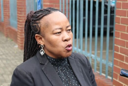 A woman with braided hair speaks passionately outdoors, wearing a black blazer and earrings. Behind her are red brick walls and metal bars.