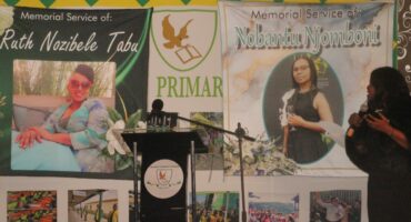 Memorial service banners for Ruth Nozibele Tabu and Nobantu Njomboni are displayed. A woman speaks at a podium with floral decorations.