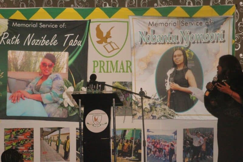 Memorial service banners for Ruth Nozibele Tabu and Nobantu Njomboni are displayed. A woman speaks at a podium with floral decorations.