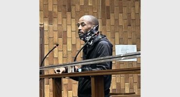 Man in a courtroom, wearing a dark jacket and patterned scarf, stands at the witness stand with a serious expression. Brick wall in the background.