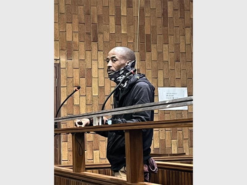 Man in a courtroom, wearing a dark jacket and patterned scarf, stands at the witness stand with a serious expression. Brick wall in the background.