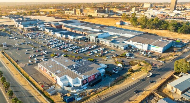 Aerial view of a large shopping center with multiple buildings and a busy parking lot filled with cars. Roads and trees surround the complex under a clear sky.
