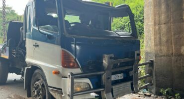 A truck with visible front-end damage is stuck under a low concrete bridge. The surroundings are wet, and green foliage is visible in the background.