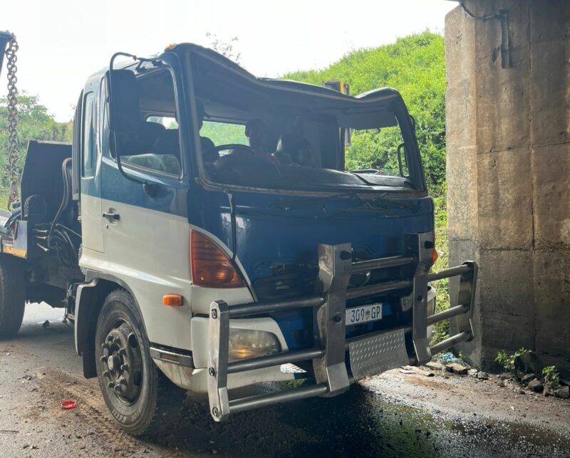 A truck with visible front-end damage is stuck under a low concrete bridge. The surroundings are wet, and green foliage is visible in the background.