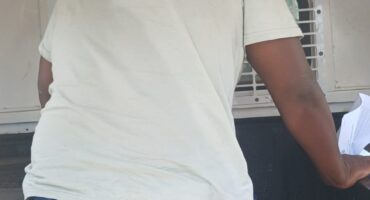 A person kneels inside a white vehicle, wearing a light shirt and blue pants. The space appears cramped, with a barred window behind them, conveying tension.