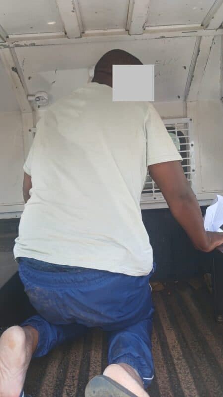 A person kneels inside a white vehicle, wearing a light shirt and blue pants. The space appears cramped, with a barred window behind them, conveying tension.