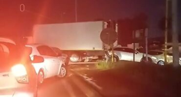 A nighttime traffic scene shows cars halted behind a truck blocking an intersection. Red taillights illuminate the road, conveying a tense and congested mood.