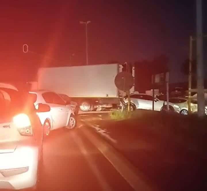A nighttime traffic scene shows cars halted behind a truck blocking an intersection. Red taillights illuminate the road, conveying a tense and congested mood.