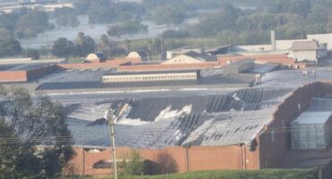 Aerial view of Festival Mall building complex with rusted metal roofs and brick walls. Background shows cooling towers and trees in a misty, rural setting.