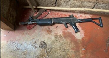 A black rifle lies on a wet, textured concrete floor beside a red broom and a wooden stick. The scene conveys tension and neglect.