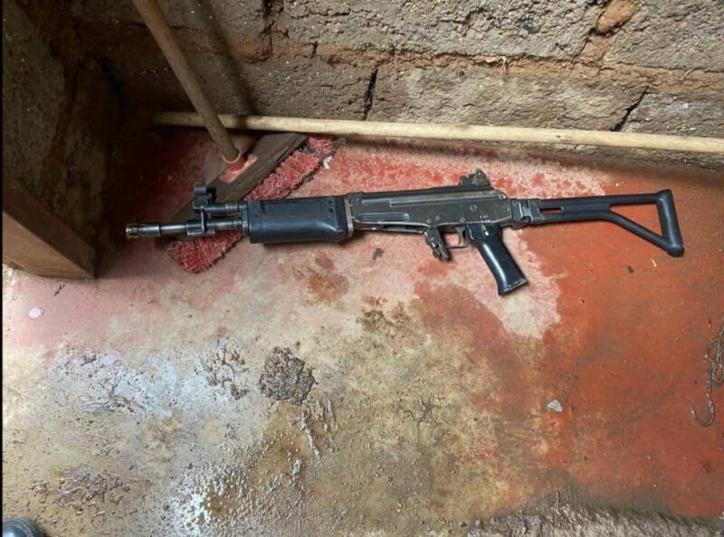 A black rifle lies on a wet, textured concrete floor beside a red broom and a wooden stick. The scene conveys tension and neglect.
