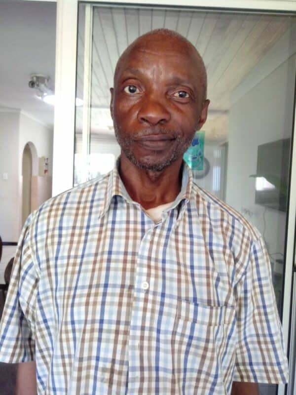A man in a plaid shirt stands indoors, near a glass door. He has a calm expression, with a neutral backdrop, conveying a quiet, homely atmosphere.