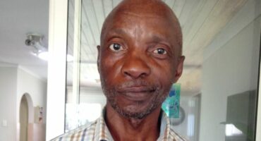 A man in a plaid shirt stands indoors, near a glass door. He has a calm expression, with a neutral backdrop, conveying a quiet, homely atmosphere.