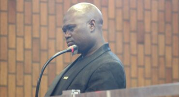 A man in a black suit stands at a microphone in a wood-paneled room. He appears serious and focused, possibly speaking or giving a testimony.