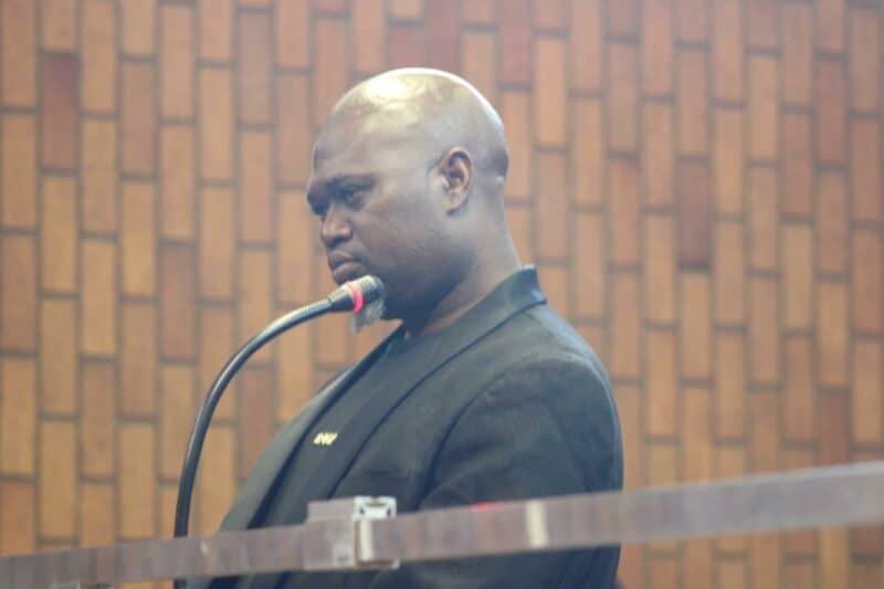 A man in a black suit stands at a microphone in a wood-paneled room. He appears serious and focused, possibly speaking or giving a testimony.