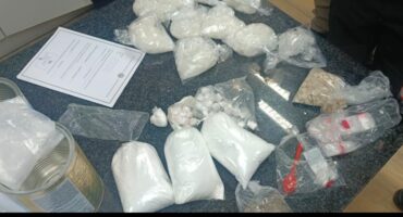 Bags of white powder and plastic-covered bundles are spread across a dark surface, suggesting a police evidence display. A paper with text is visible.